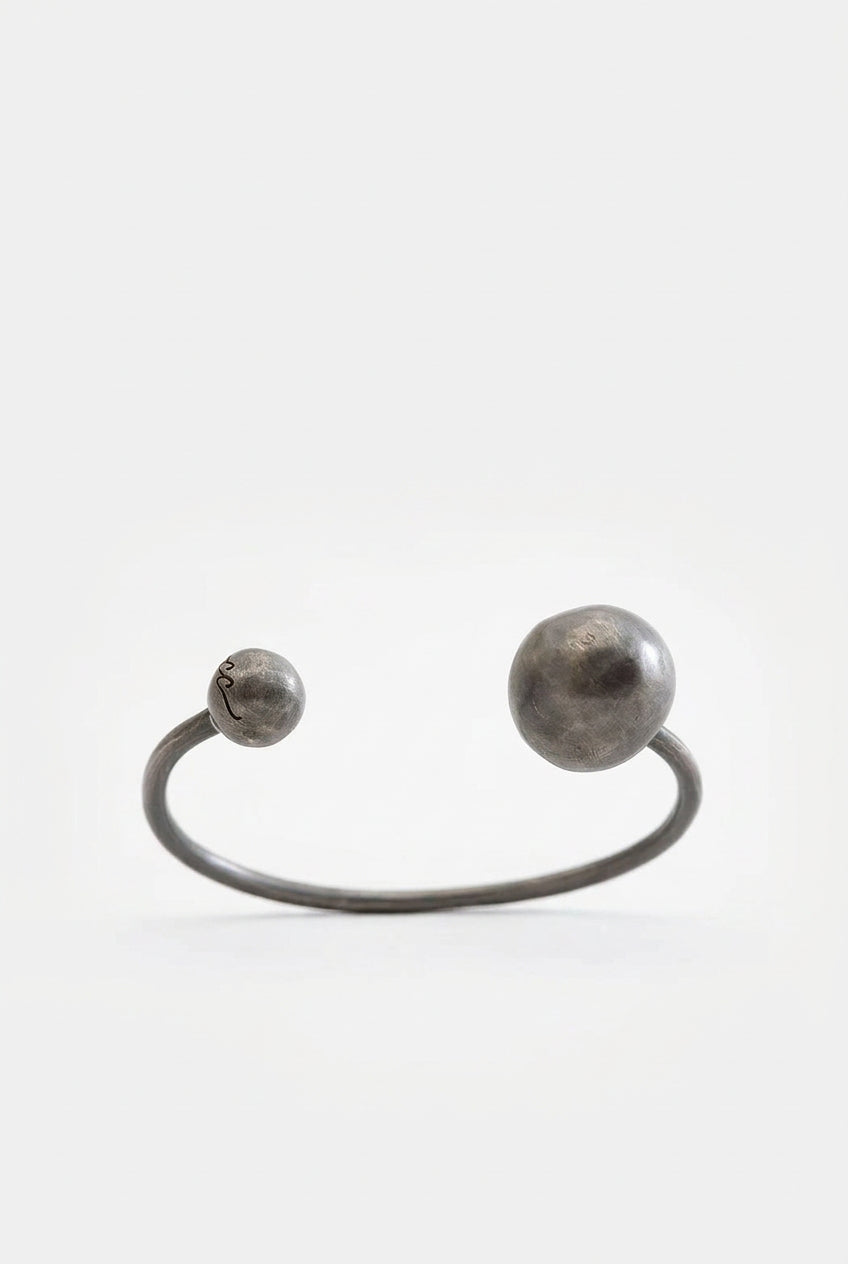 A minimalist open cuff bracelet made of textured dark gray metal, featuring two spherical ends, one larger and one smaller, set against a plain light gray background.