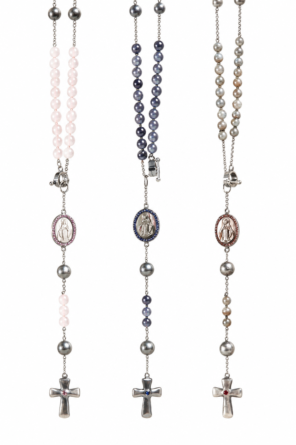 Three rosary necklaces with silver-tone chains and crosses are displayed vertically against a white background. The left rosary features light pink beads, the middle features dark blue beads, and the right features gray beads, each with corresponding medallions and a crucifix.