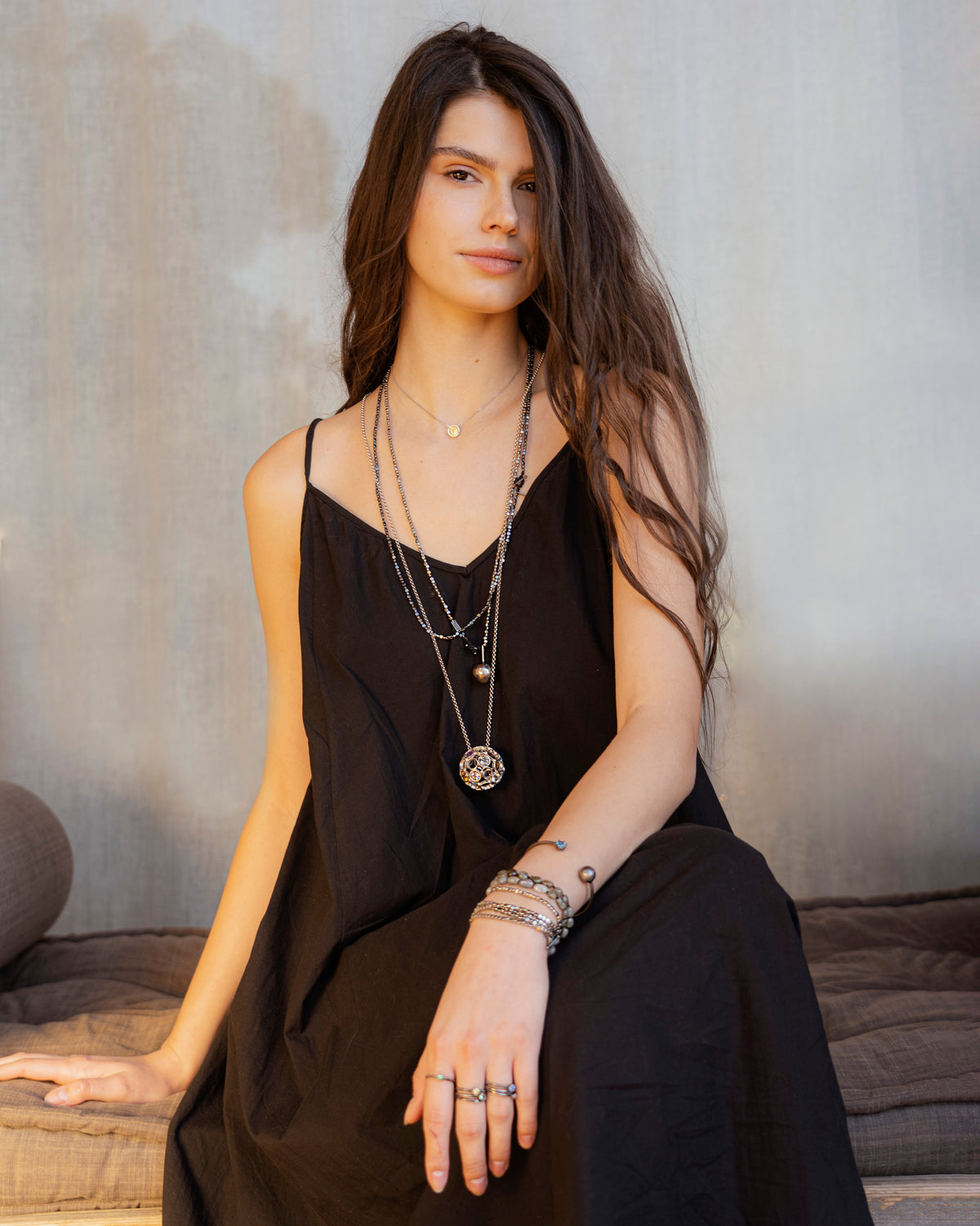 Woman wearing a black dress with layered necklaces and bracelets against a neutral background