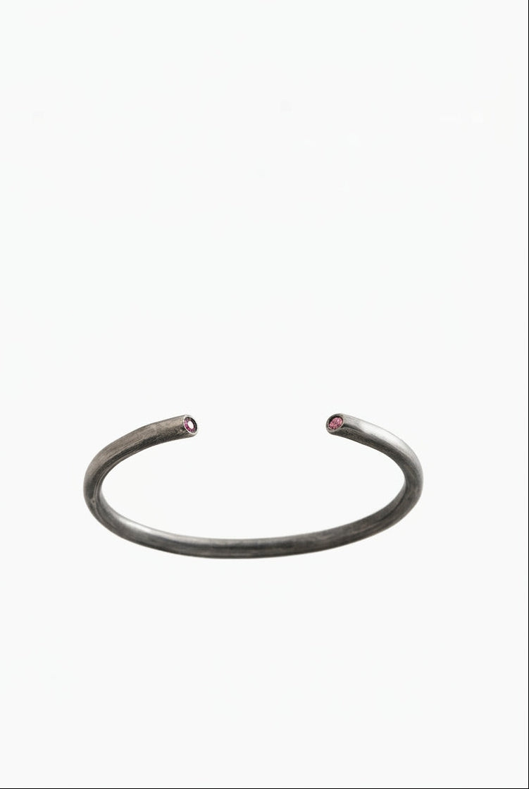 A minimalist open cuff bangle bracelet in a dark, textured gunmetal gray metal, featuring tiny red gemstone accents at each end, set against a solid white background.