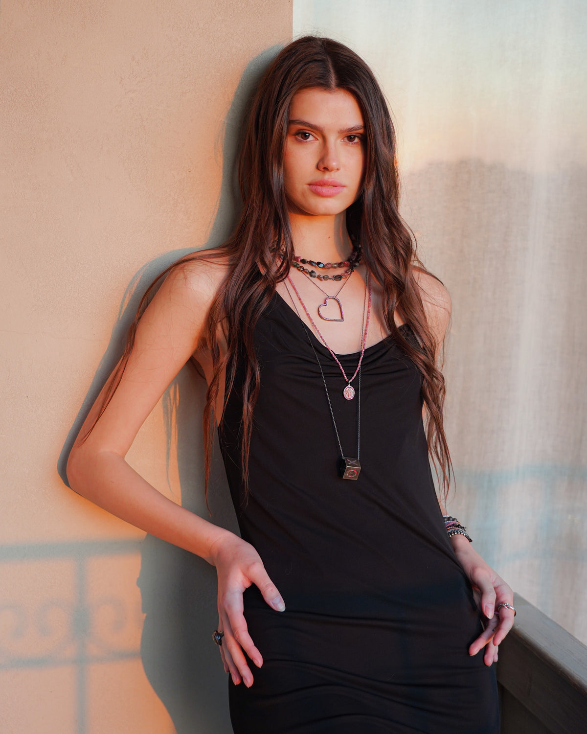 Woman wearing a black sleeveless top with layered necklaces against a neutral wall.